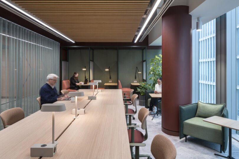 People working at a long wooden table in a brightly lit office with large windows and plants.