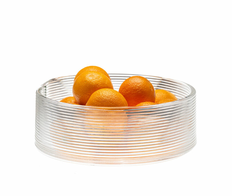 A clear, ribbed 3D printed glass bowl filled with fresh oranges.