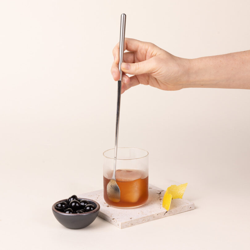 A hand using the metal stirring spoon to mix a drink in a glass with ice and garnishes.