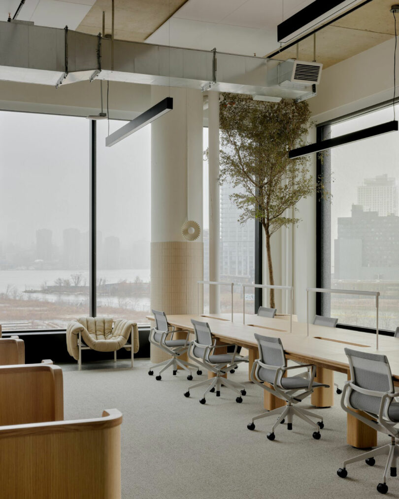 Large open-plan office space featuring long wooden communal tables and ergonomic gray chairs with city views.