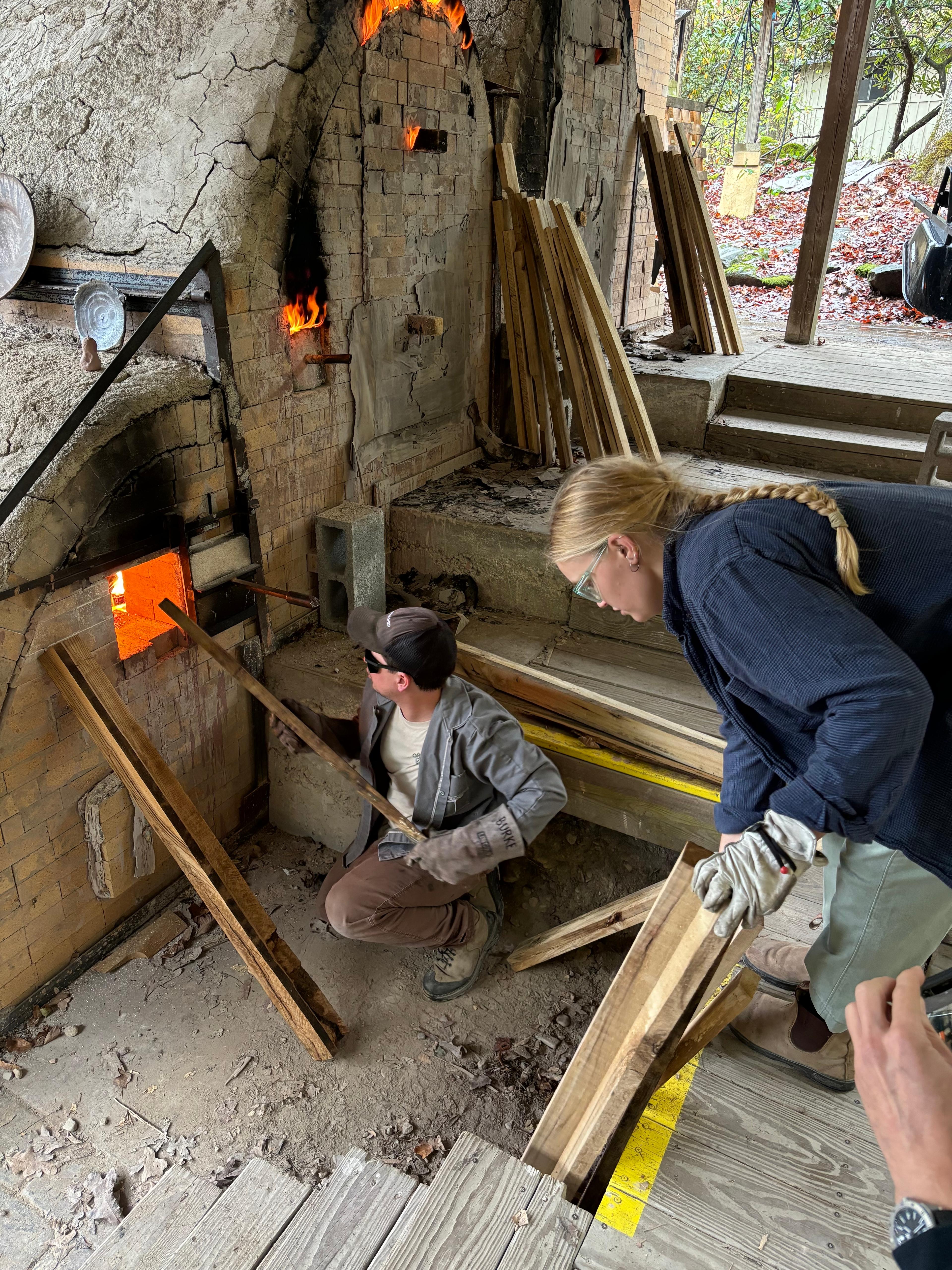 A ceramic artist stoking the fire box of a wood kiln with long pieces of firewood.