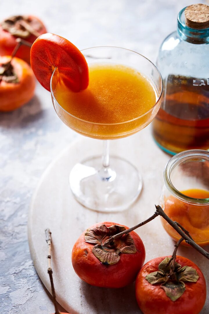 An amber-colored bourbon cocktail garnished with a slice of persimmon in a heavy-bottomed glass.