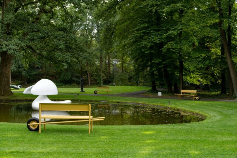 Several yellow benches scattered across a lawn near a pond with large white sculptures and tall trees.