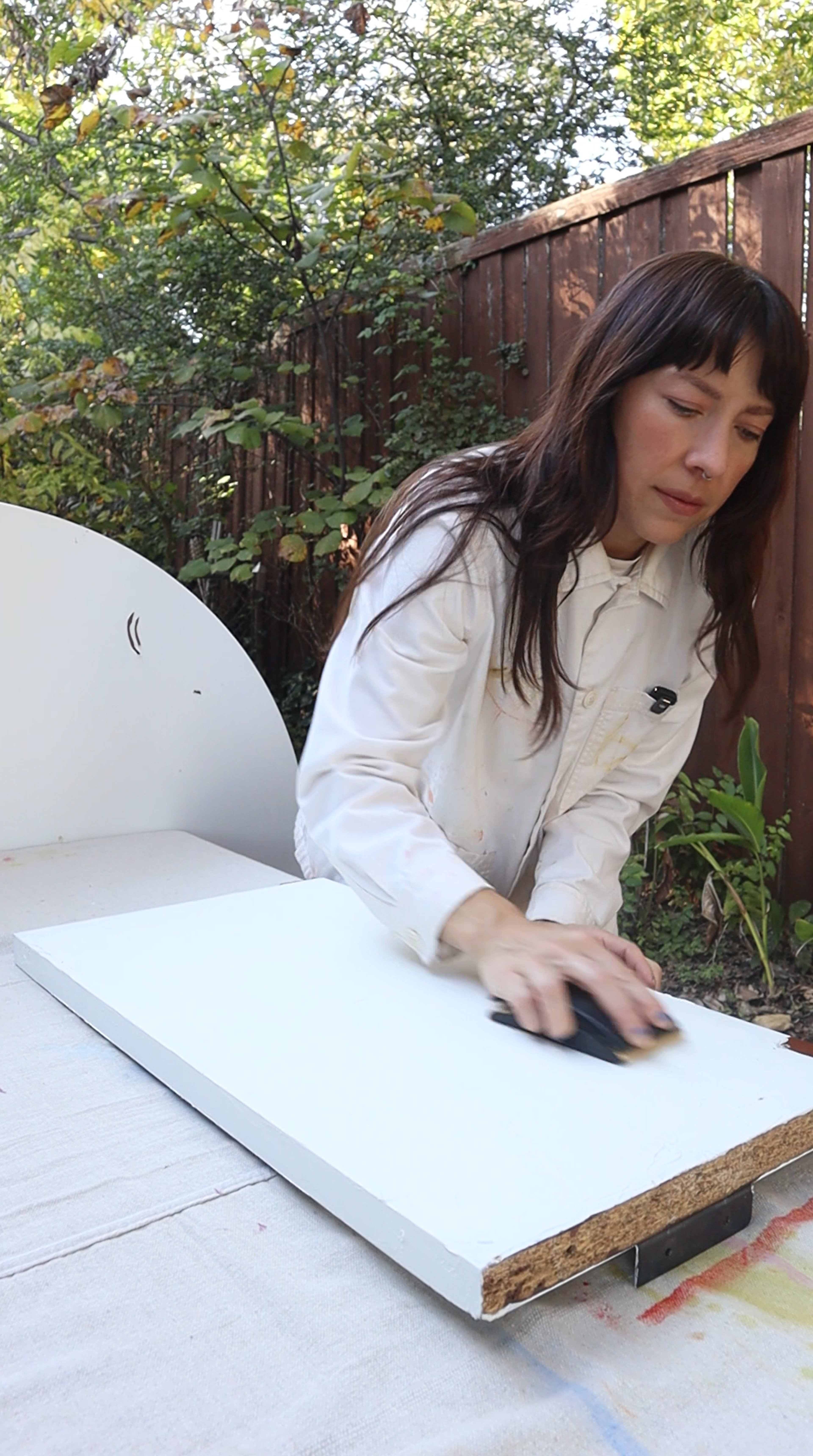 A person using a hand sander on a wooden table surface with sawdust visible.