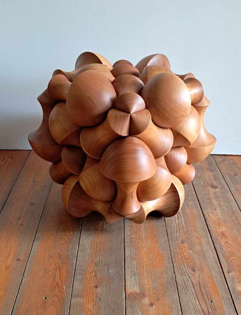 A bulbous cherry wood geometric sculpture standing against a plain white wall on a wooden floor.