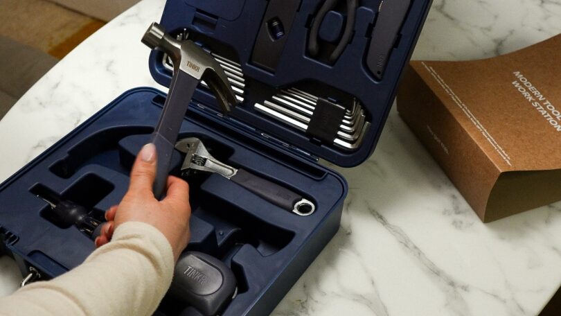 A hand reaches for a hammer inside an open blue toolkit resting on a white marble countertop.