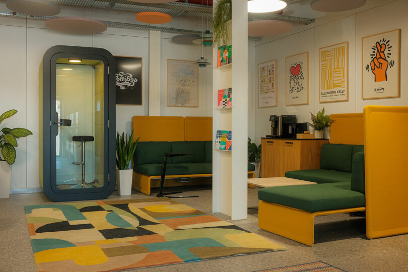 A modern office lounge featuring a private phone booth, mustard-colored seating, and decorative plants.