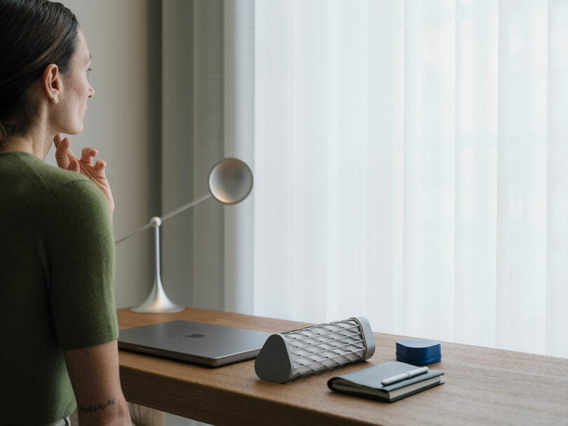 A clean, modern wooden desk featuring a laptop, notebook, and an Elio water bottle next to a window.
