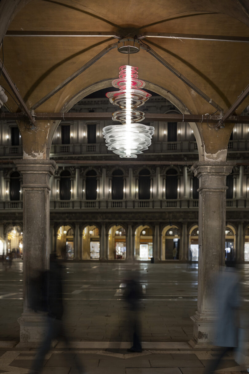 Atmospheric view of a modern pendant light in a stone archway with historic buildings in the background.