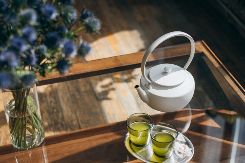 A white teapot and two cups of green tea on a glass table with blue flowers and soft shadows.