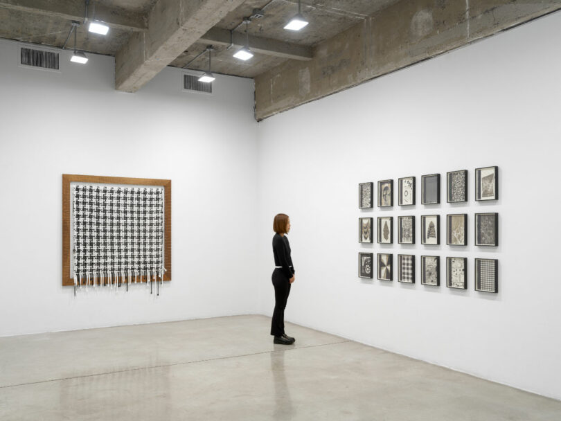 An installation view showing a framed orange painting alongside a grid of eighteen framed technical sketches on a white wall.