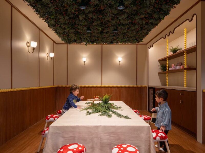 Children standing near mushroom-shaped stools in a room with a leafy ceiling installation.