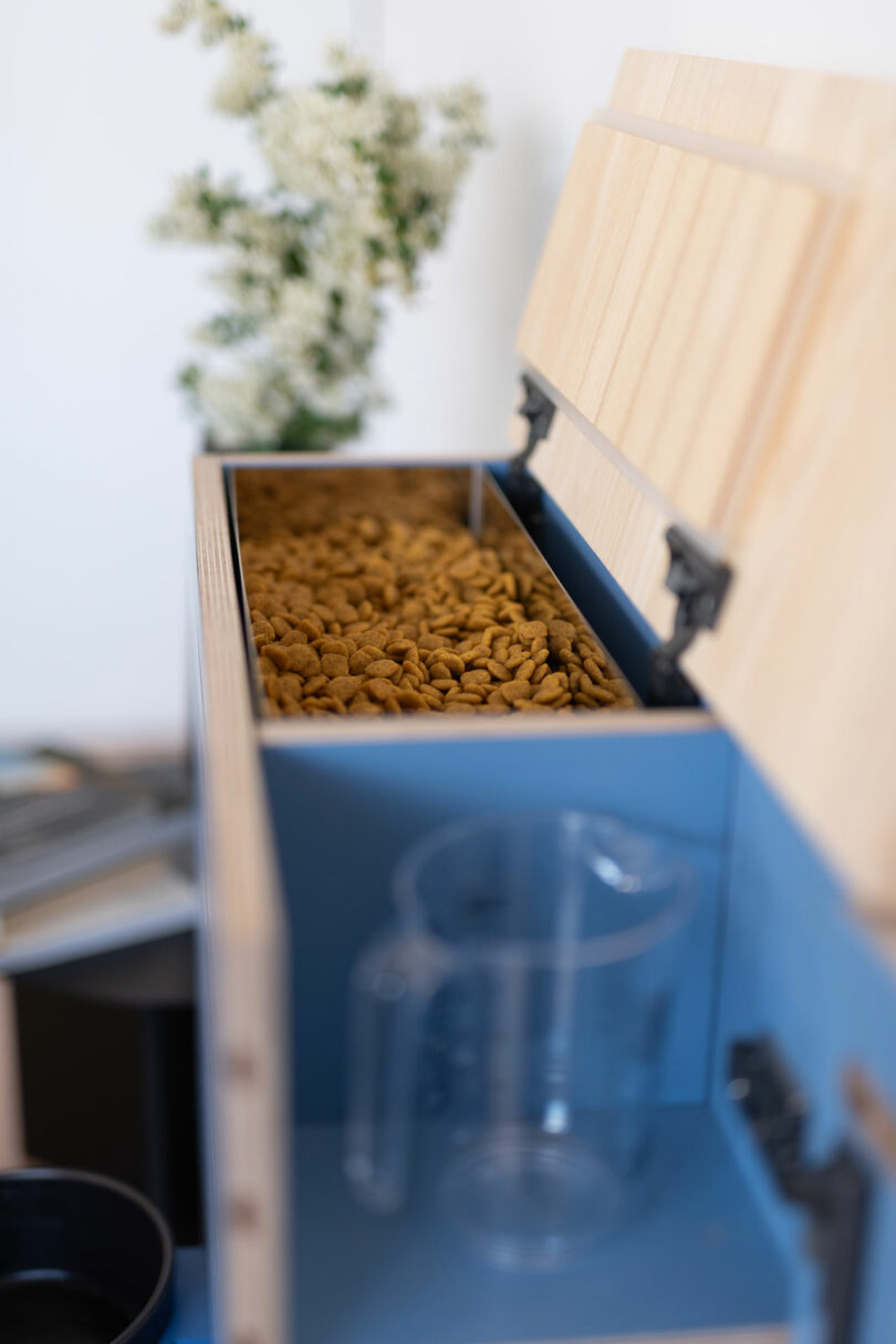 A close-up of a natural wood pet food storage box with dry kibble and a clear measuring cup.
