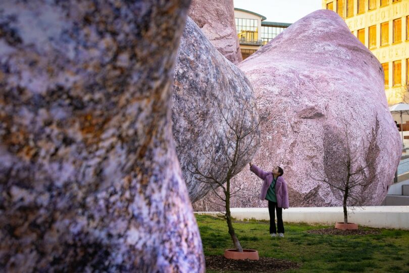 A person standing among purple-toned, textured rock sculptures in an urban plaza.