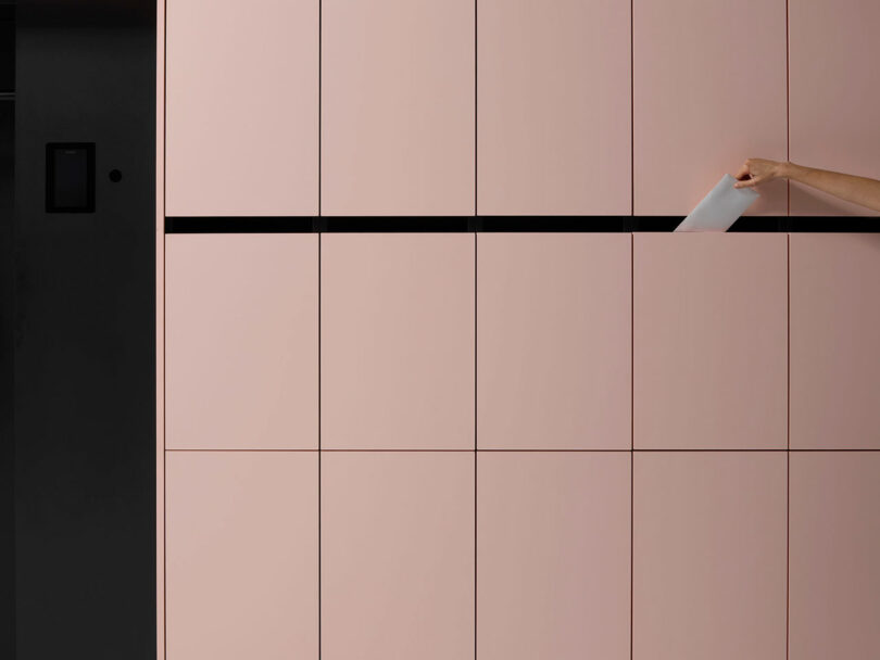 A hand inserting a white envelope into a horizontal mail slot integrated into the pink locker doors.