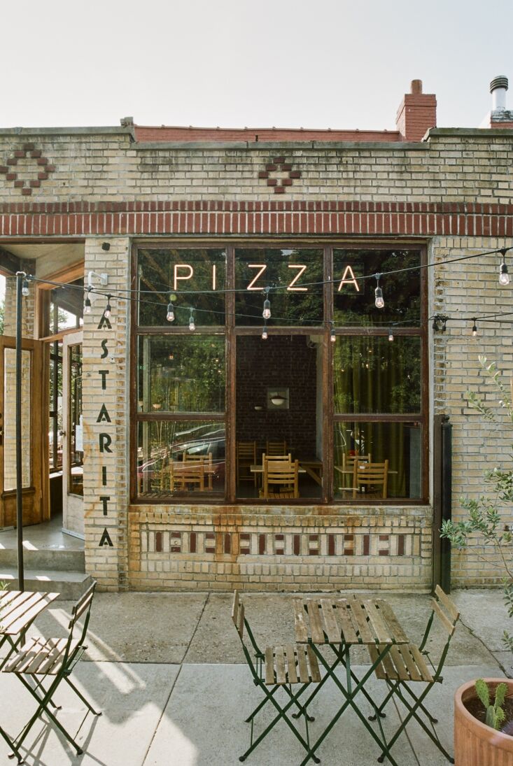 The exterior of Astarita Pizzeria at a sunny street corner in Greenwood Heights, Brooklyn, featuring yellow brick and sidewalk tables.