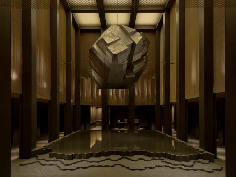 A geometric stone sculpture suspended over a dark reflection pool in a dimly lit, modern architectural space.