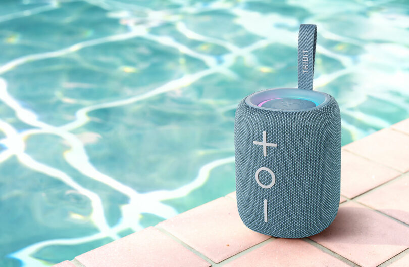A blue TRIBIT speaker sitting safely on the edge of a swimming pool with water splashing nearby.