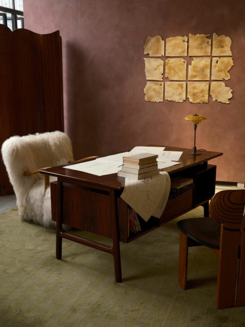 A mid-century wooden desk and fuzzy chair in a softly lit room with abstract artwork on a mauve wall.