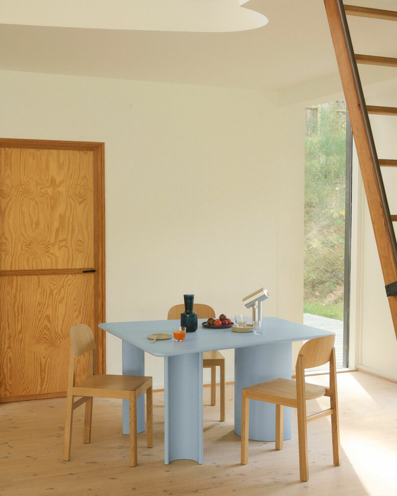 Light blue Re-norm Table in a minimalist room with large windows, wooden chairs, and indoor plants.