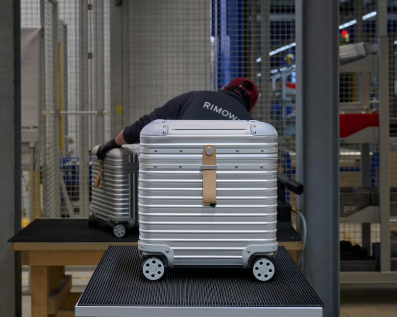 A technician in a factory environment assembling a silver grooved aluminum suitcase shell.