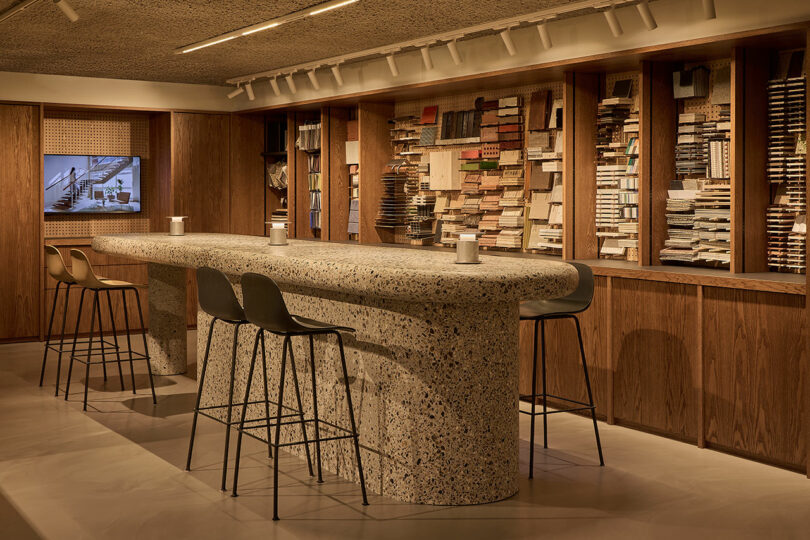 A high terrazzo-topped table with black stools in a modern showroom setting with material samples in the background.
