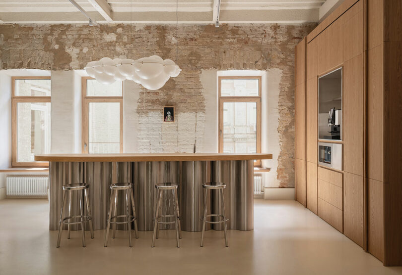 A modern office kitchen with a long wooden island, metal stools, and an exposed brick wall.