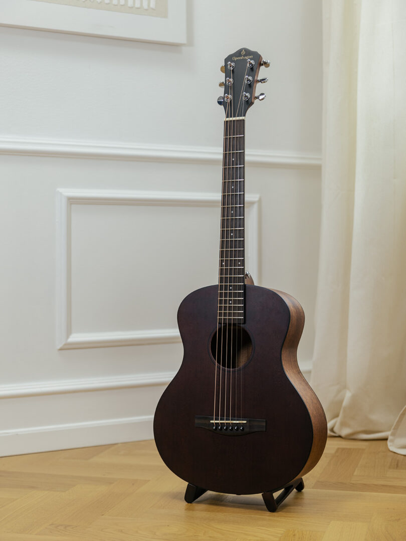 A dark brown acoustic guitar standing on a small wooden stand in a clean white room with hardwood floors.
