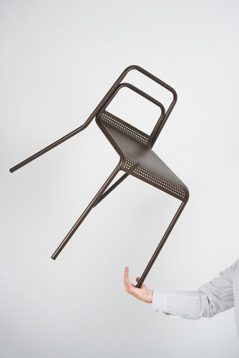 A person balancing a lightweight perforated metal chair on their palm against a white background.