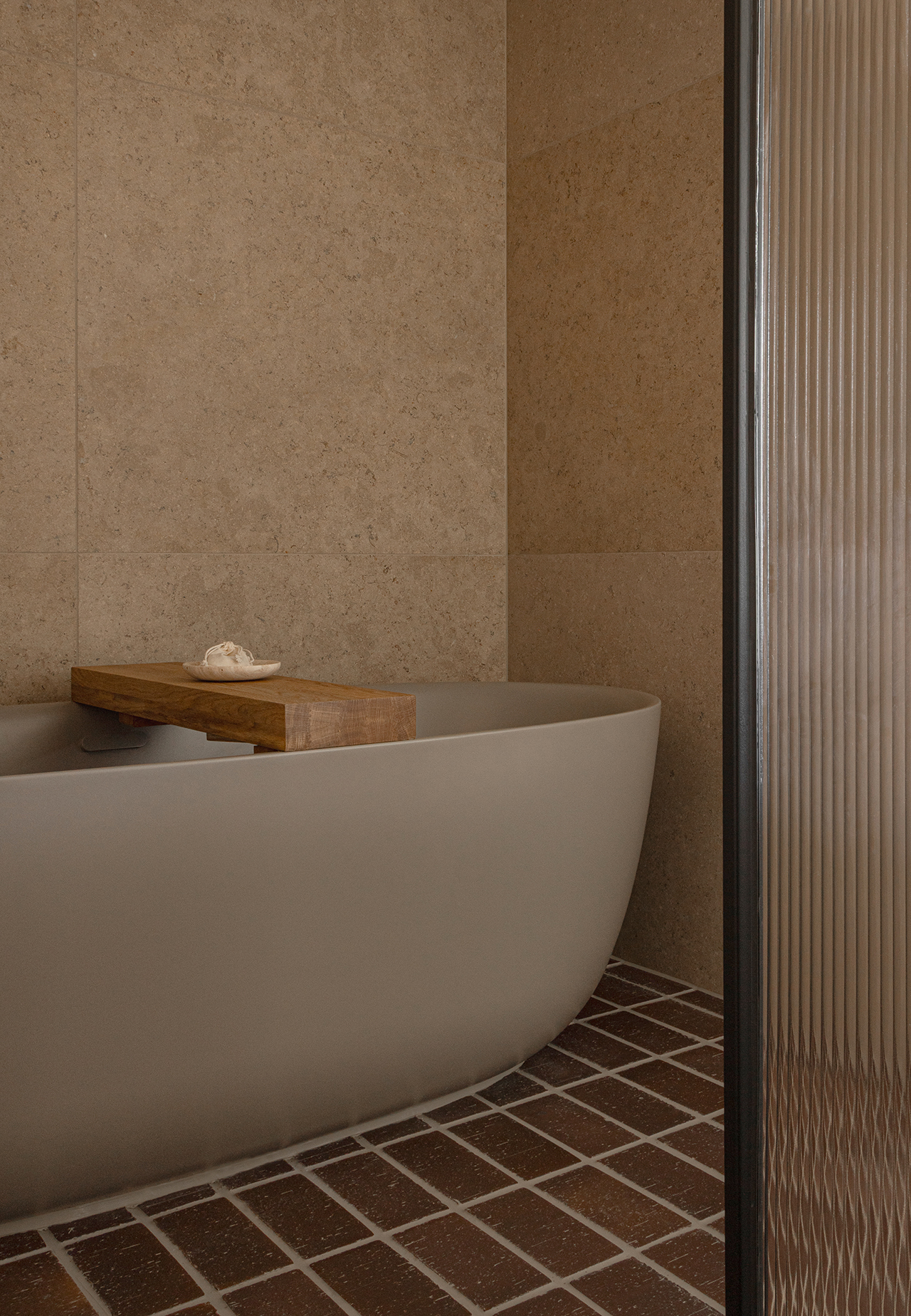 A minimalist bathroom with a freestanding white bathtub, beige stone walls, and warm wooden accents.