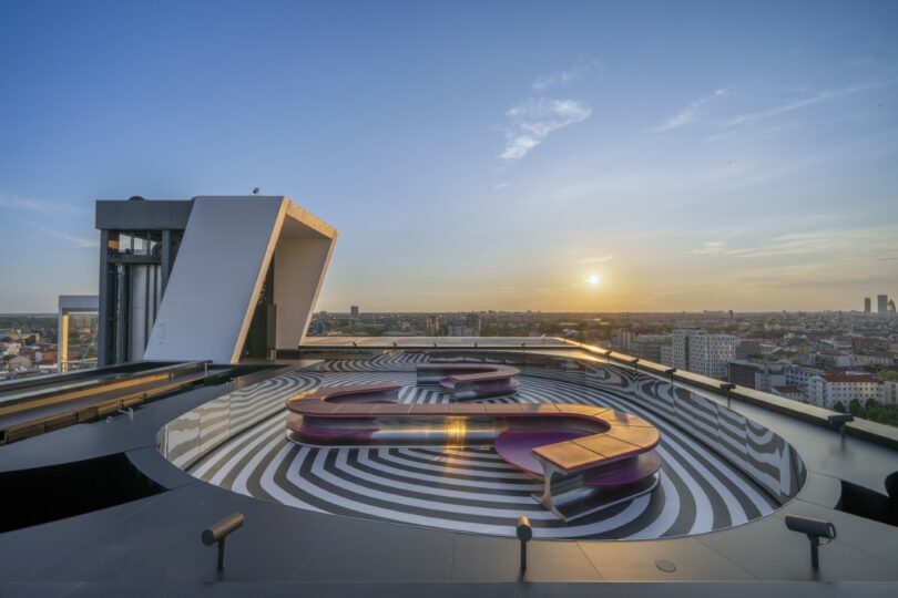 The rooftop terrace at sunset featuring purple sculptural benches and the glowing city lights in the background.