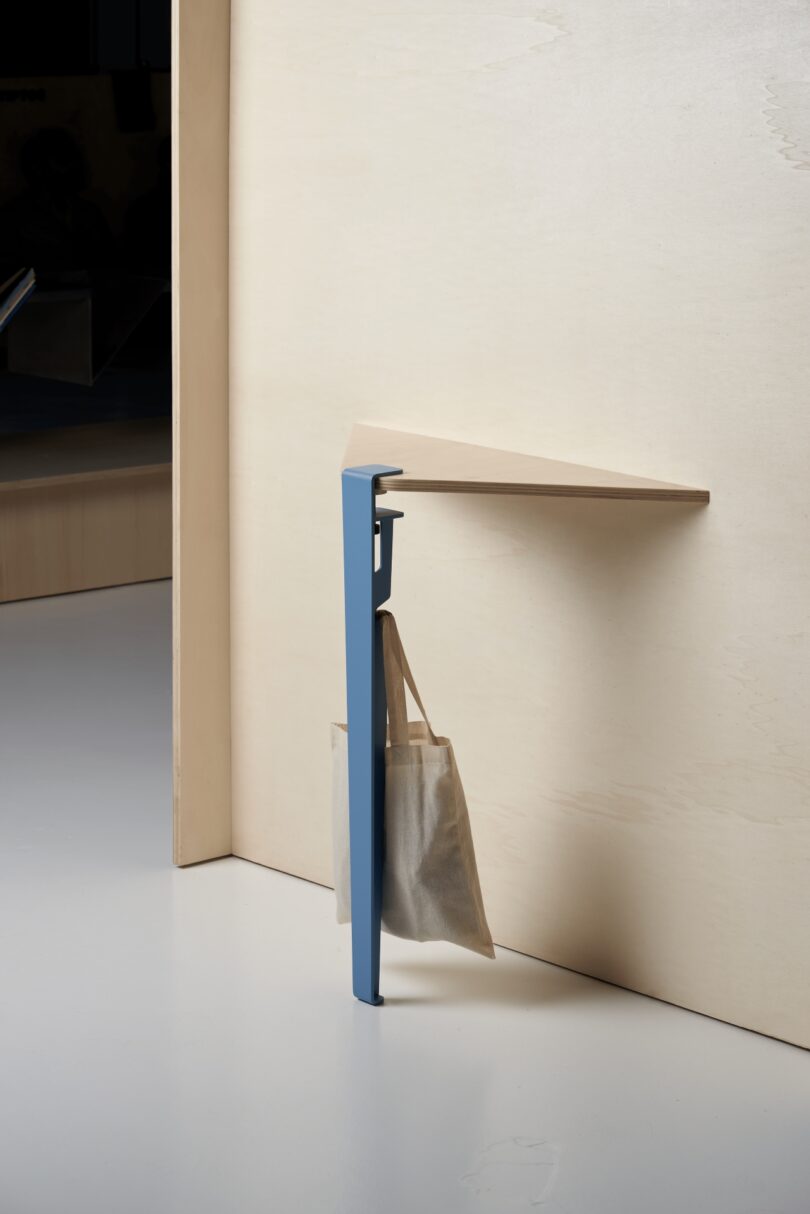 A wooden corner shelf supported by a blue metal Tiptoe leg with a built-in hook.
