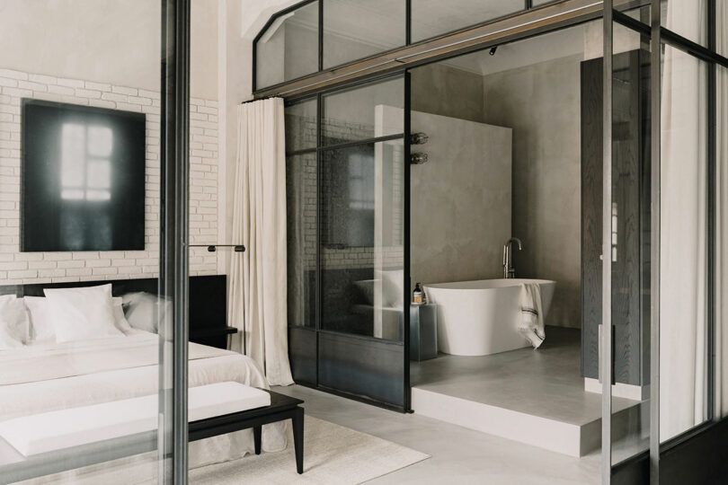 Modern bedroom and bathroom separated by a glass divider, featuring neutral tones and a freestanding tub.