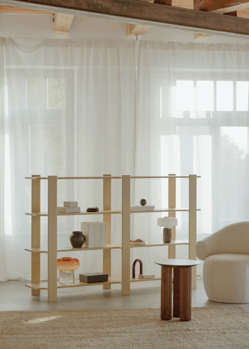 A beige open-shelf bookcase in a minimalist living room with a white armchair and sheer curtains.
