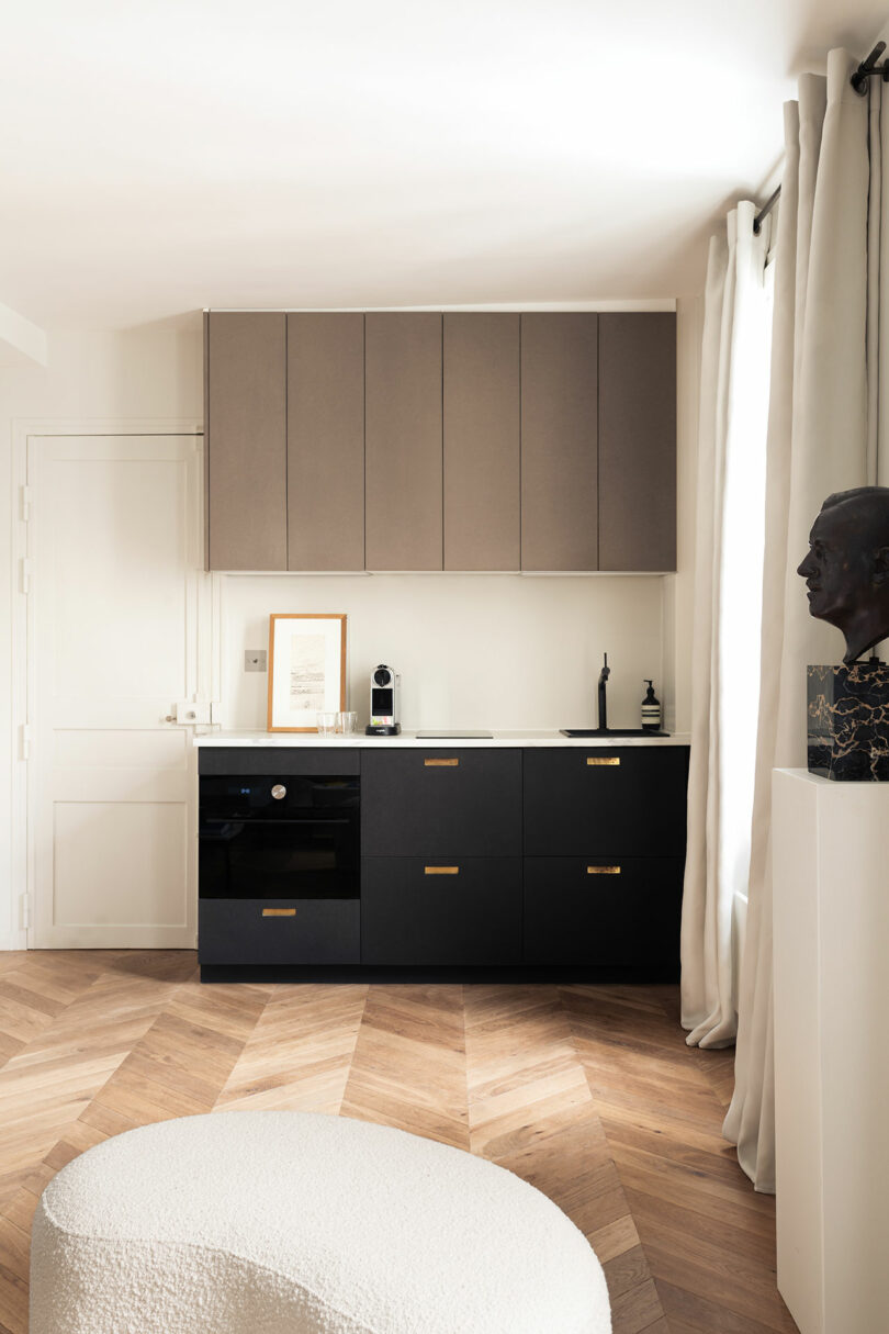 A minimalist kitchen area with black and wood cabinetry, a decorative bust, and classic wood herringbone flooring.