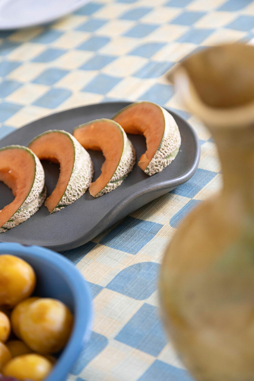 Slices of fruit on a gray plate next to a blue bowl on a checkered tablecloth.