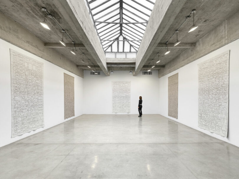 A sunlit white-walled art gallery featuring five long vertical white tapestries hanging from the ceiling.
