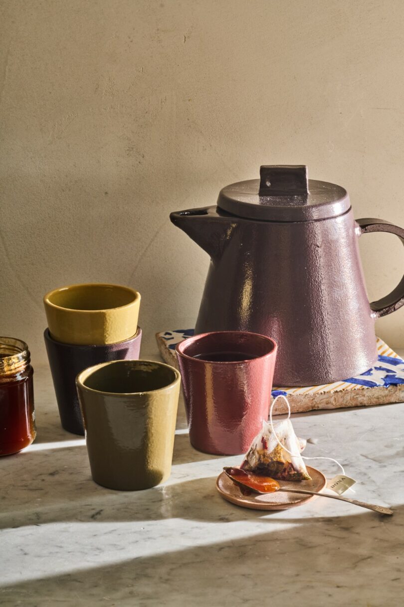 A ceramic teapot and glazed cups in an earthy tone resting on a marble surface.