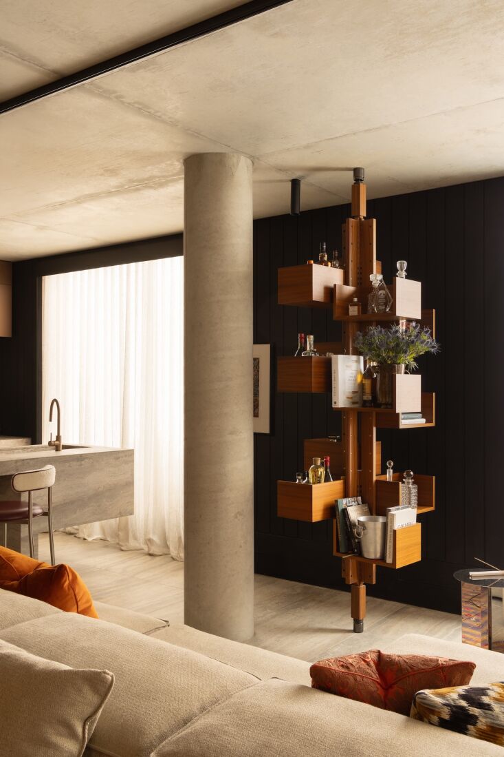 A mid-century modern wooden bookcase used as a bar standing against a structural concrete column and ceiling.