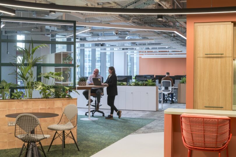 Two people working on a laptop at a modern standing desk within a bright, tech-forward office.