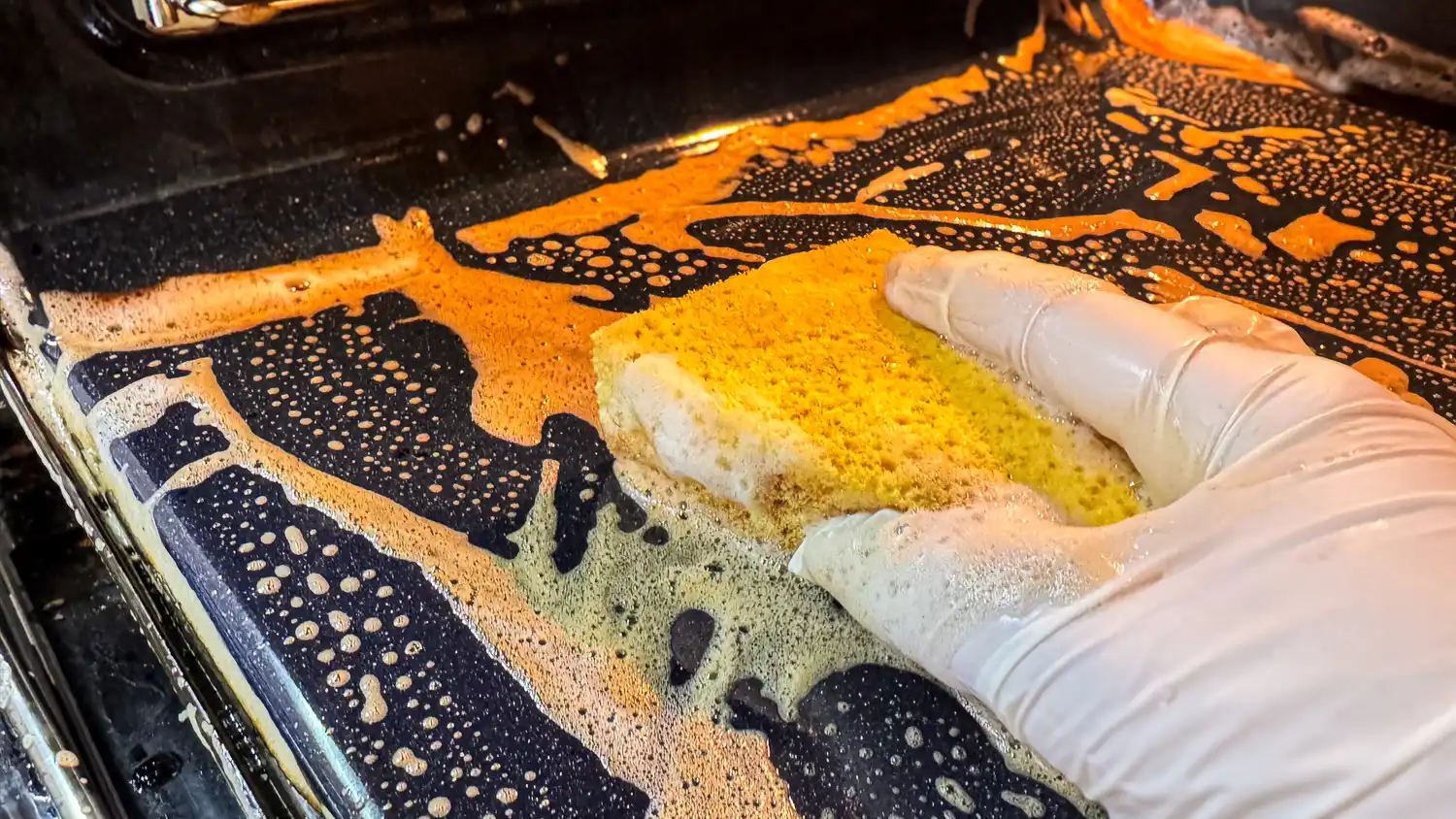 A gloved hand using a sponge to wipe down the interior black surface of an oven.