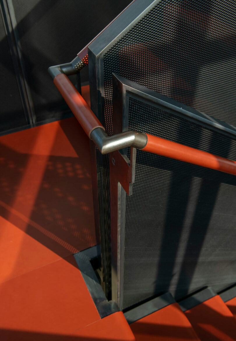 Close-up of a perforated metal staircase with a red handrail casting geometric shadows.