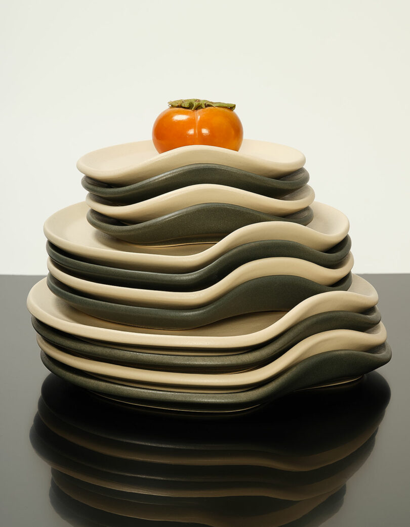 A stack of wavy-edged plates in beige and dark green with a persimmon on top.