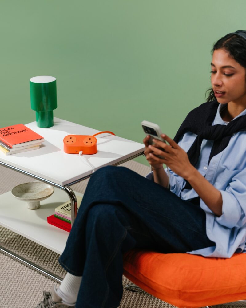A woman using her phone in a cozy living room with a green lamp and an orange charging hub on a nearby side table.