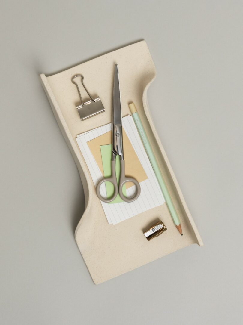 A close-up of a beige ceramic tray holding scissors, a pencil, and a stack of cards.