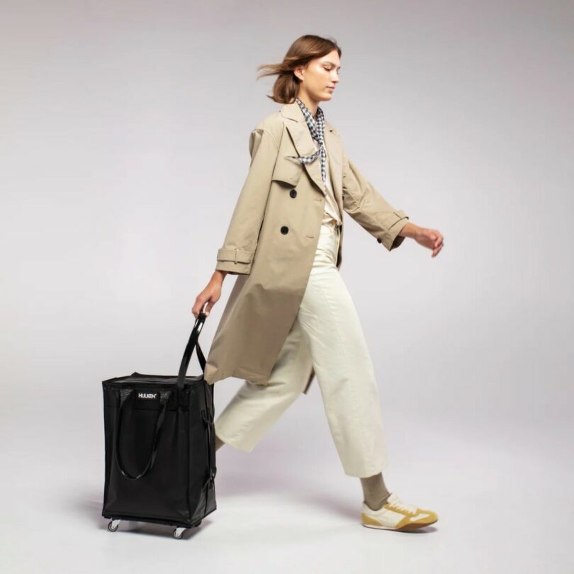 A woman in a beige trench coat pulling a black multi-wheeled rolling tote bag.