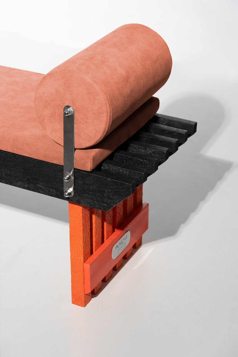 A Torii bench with an orange cushion and a cylindrical bolster pillow, featuring a metallic brand plate.