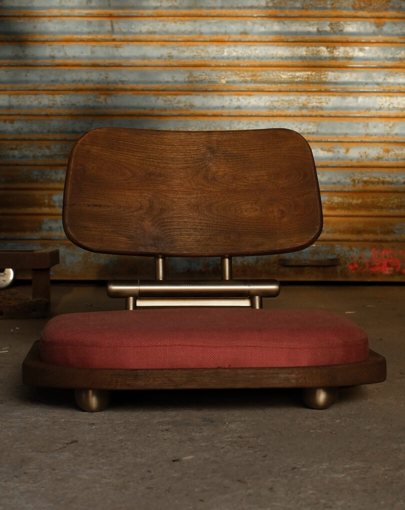 A low-profile wooden chair with a deep red cushion set against an industrial corrugated metal backdrop.