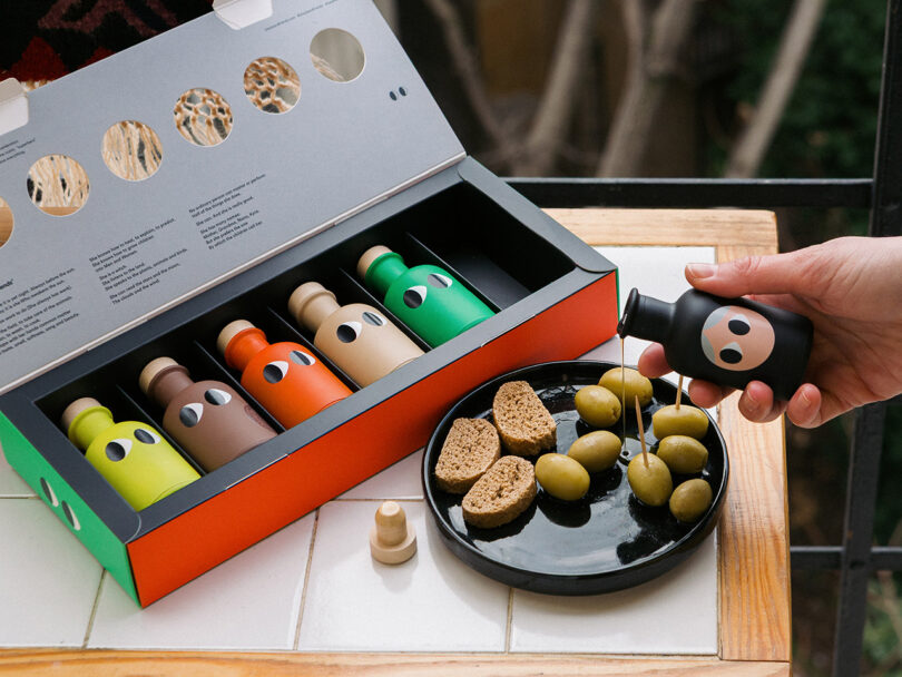 A hand pouring oil from a small black bottle onto bread and olives, with a gift box of colorful bottles nearby.