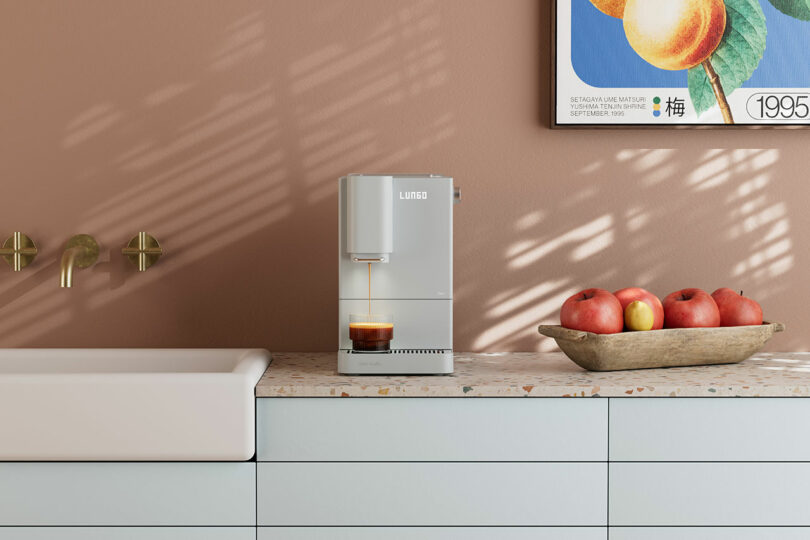 A black Terra Kaffe machine brewing coffee on a trendy terrazzo countertop next to a bowl of red apples.